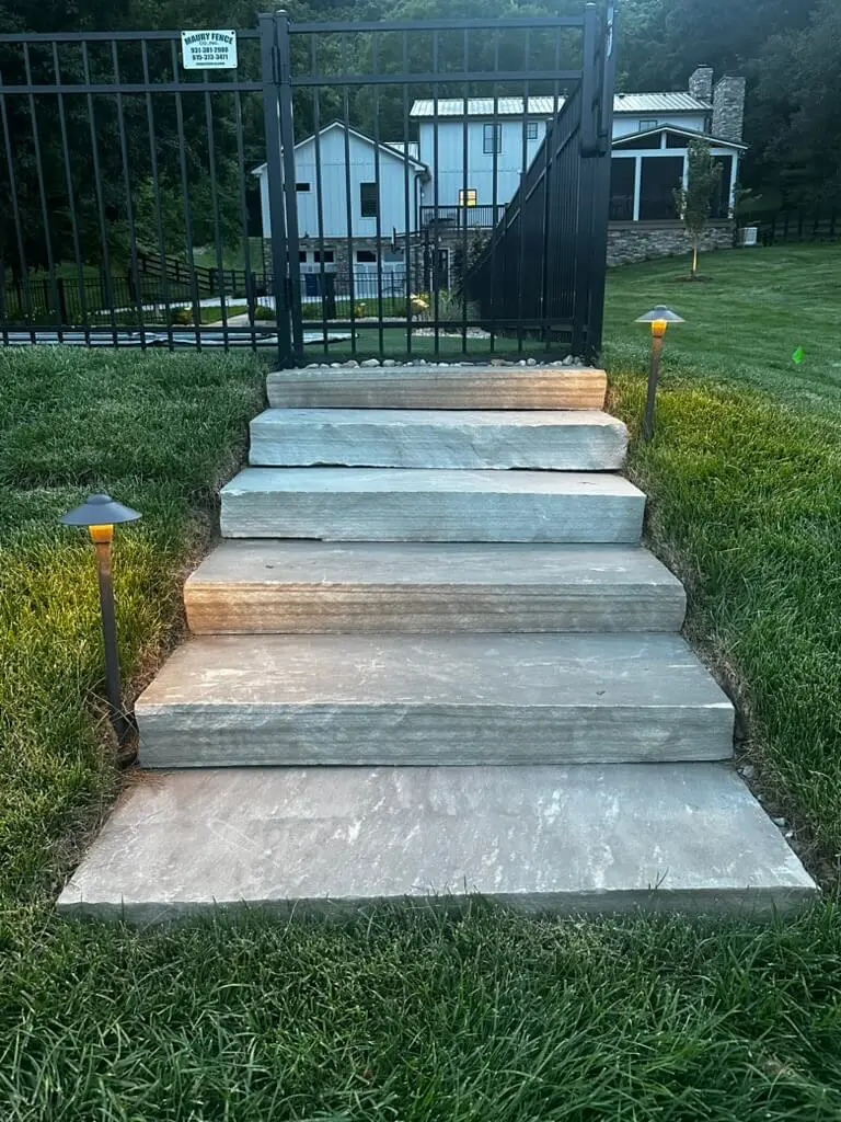 Stone steps lead up to a black metal gate, flanked by grass and two pathway lights. A house and trees are visible in the background.