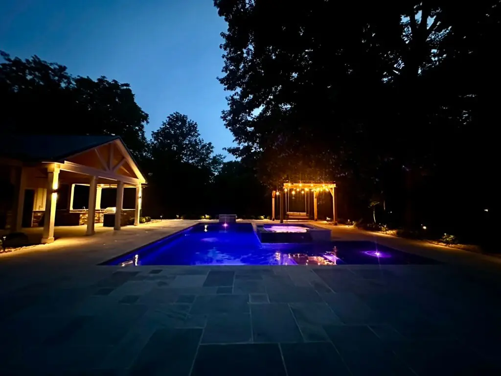 A well-lit pool area at dusk with surrounding trees. The pool has glowing lights and a nearby pergola is illuminated by string lights.