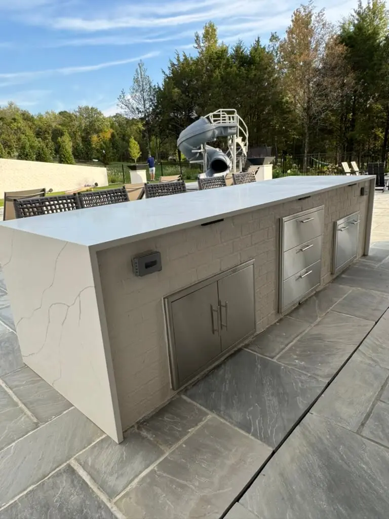 Outdoor kitchen with a marble countertop, stainless steel appliances, and a backdrop of trees and a slide.