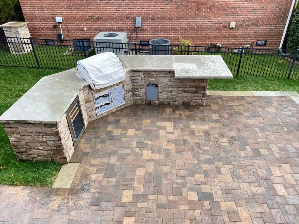 Outdoor kitchen with stone countertops and brick structure, featuring a covered grill, storage cabinets, and a sink. The area is on a paved patio with a view of a brick wall and fenced yard.