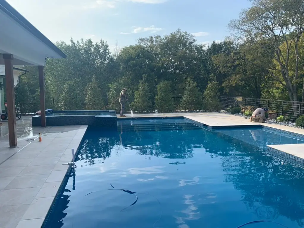 Two people work around a modern outdoor pool surrounded by trees and a tiled patio under a clear sky.