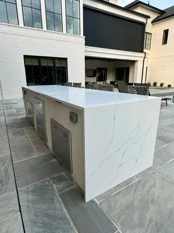 Outdoor kitchen island with a white marble countertop, stainless steel appliances, and seating area on a stone patio in front of a modern building.