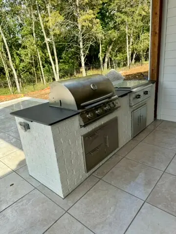 Outdoor kitchen with a stainless steel grill and storage cabinets on a tiled patio, surrounded by trees.