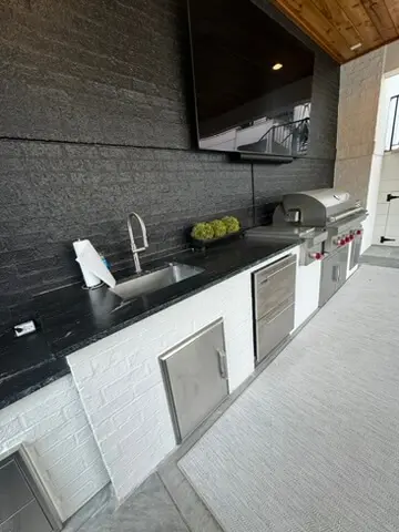 Outdoor kitchen with black countertop, sink, grill, and cabinets. A large TV is mounted above, and a plant decorates the countertop.