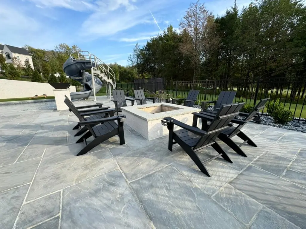 A patio with a central fire pit and black chairs, surrounded by trees. A slide is visible in the background.