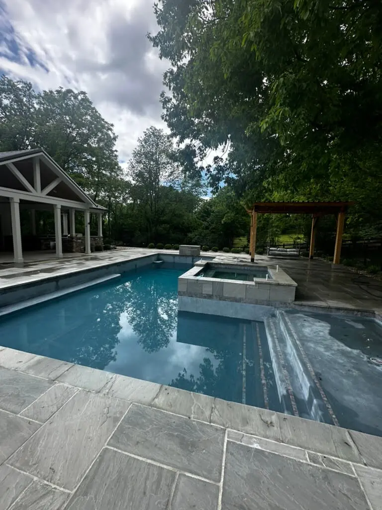 Outdoor pool with a hot tub surrounded by stone tiles, next to a wooden pergola and a pavilion, all set against a backdrop of trees and a partly cloudy sky.