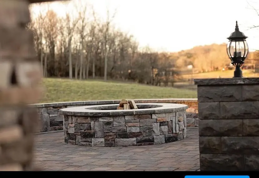 A stone fire pit stands on a paved patio, surrounded by a low stone wall. A lamppost is visible on the right. Leafless trees and a grassy field are in the background.