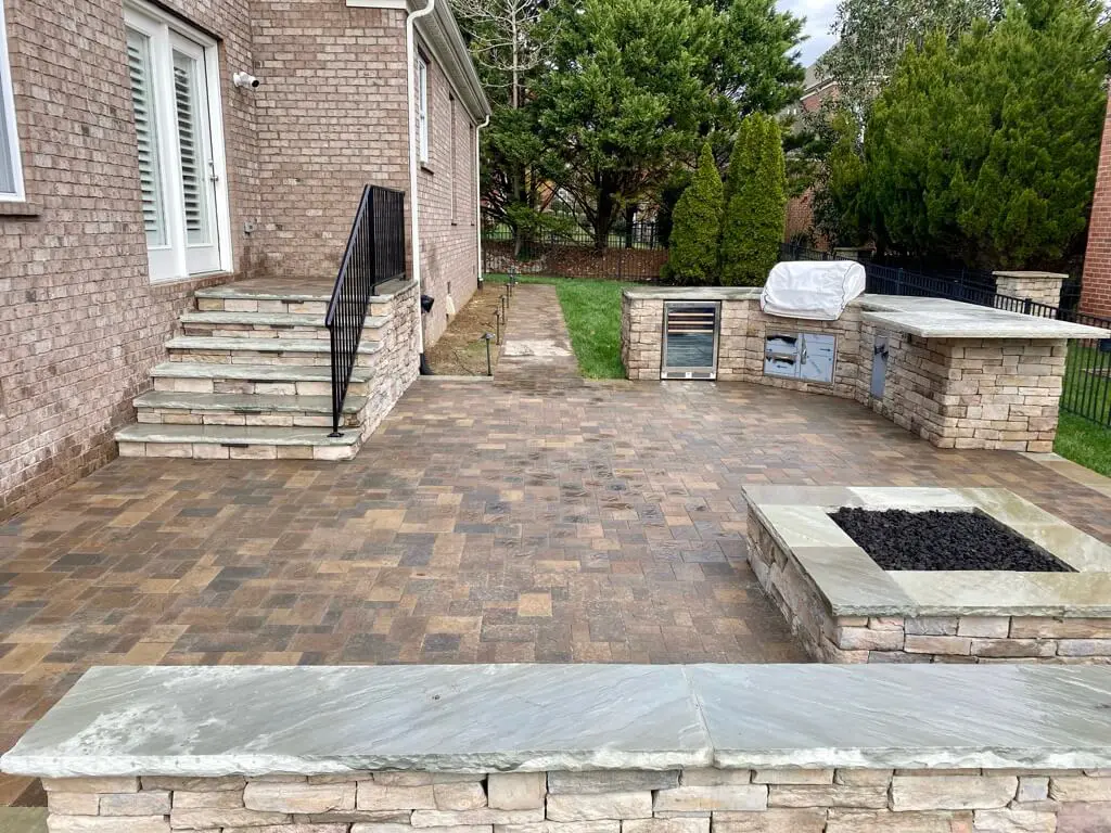 Outdoor patio with stone flooring, a built-in grill, counter space, and a fire pit. Stone steps lead to the house, and there are bushes and trees along the fence.