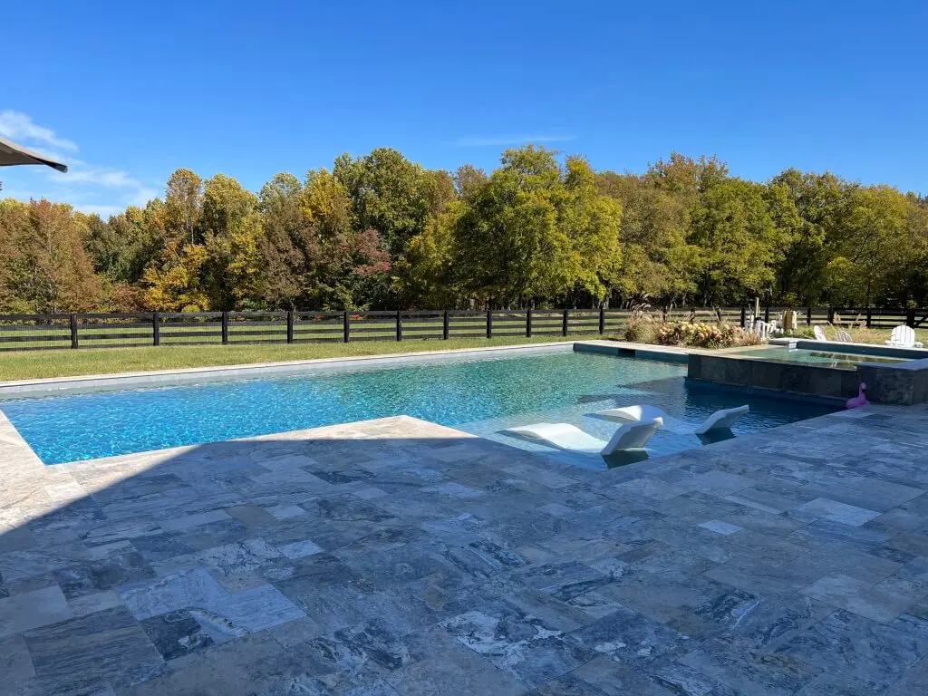 A large outdoor pool with surrounding stone tiles, two submerged lounge chairs, adjacent hot tub, and a backdrop of trees and a fence under a clear blue sky.