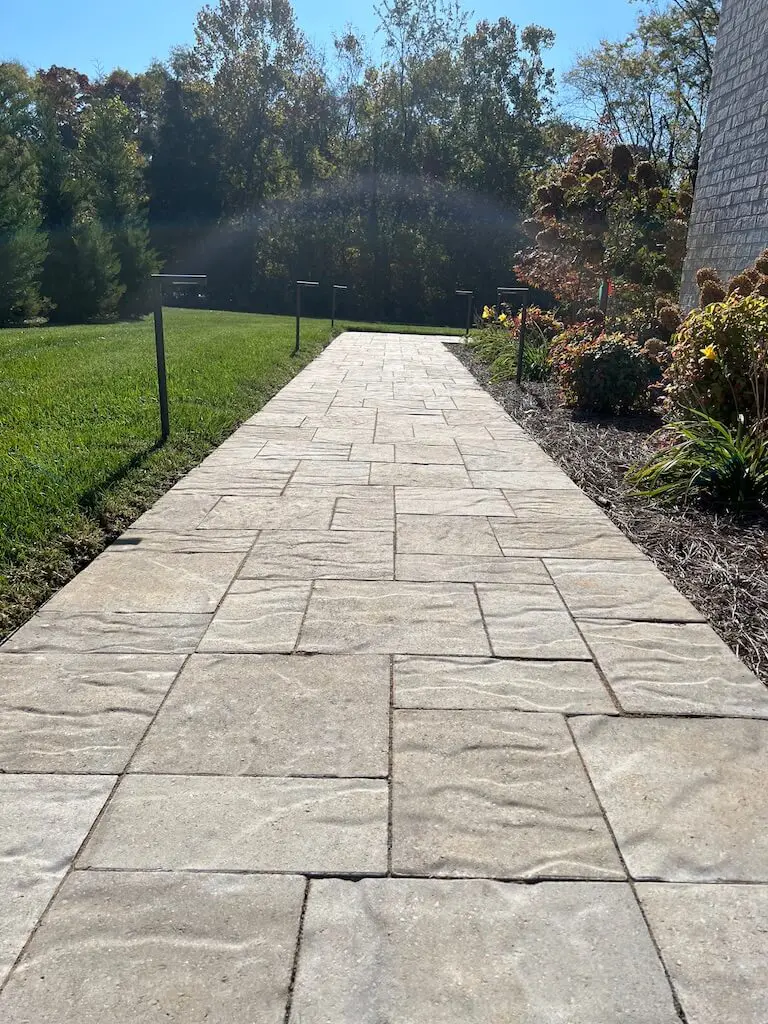 A paved walkway leads through a garden with plants and trees under a clear sky.