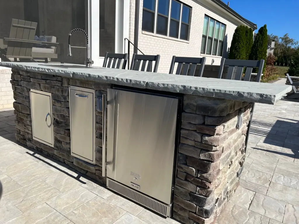 Outdoor kitchen with stone countertop, built-in grill, refrigerator, and storage under a clear blue sky.