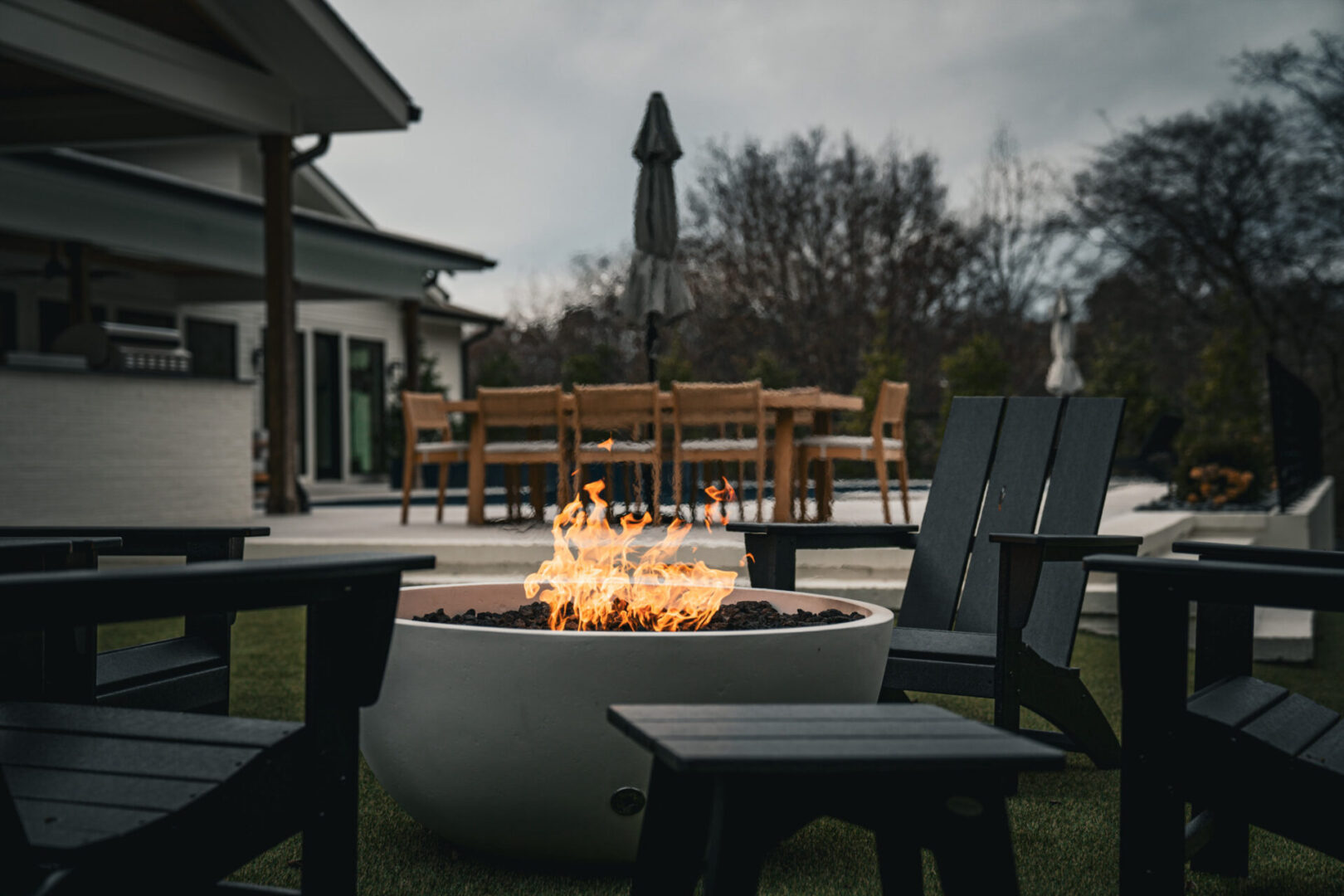 Outdoor fire pit with chairs and a dining table in the background during dusk.
