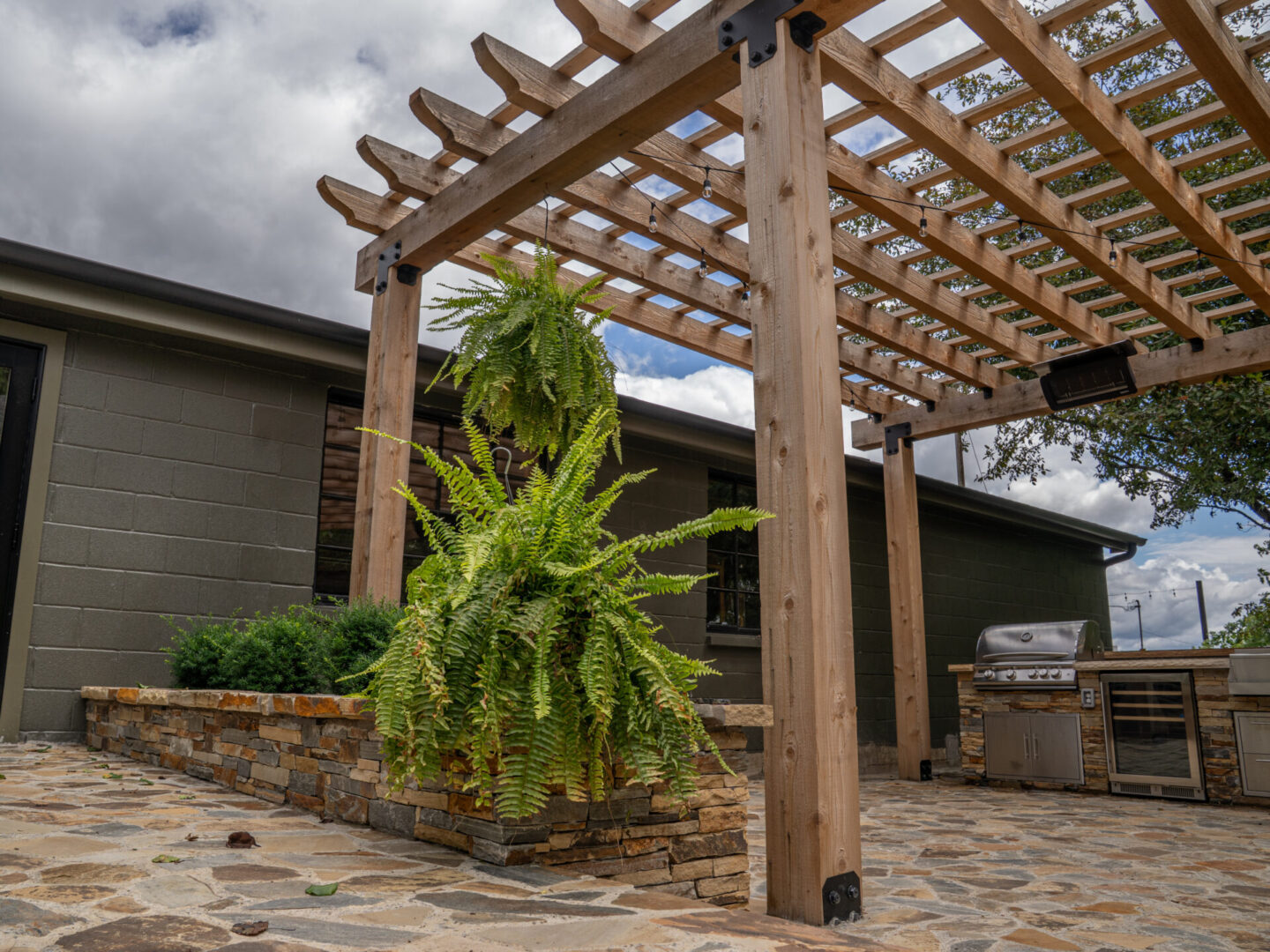 Backyard patio with wooden pergola and lush greenery under a cloudy sky.