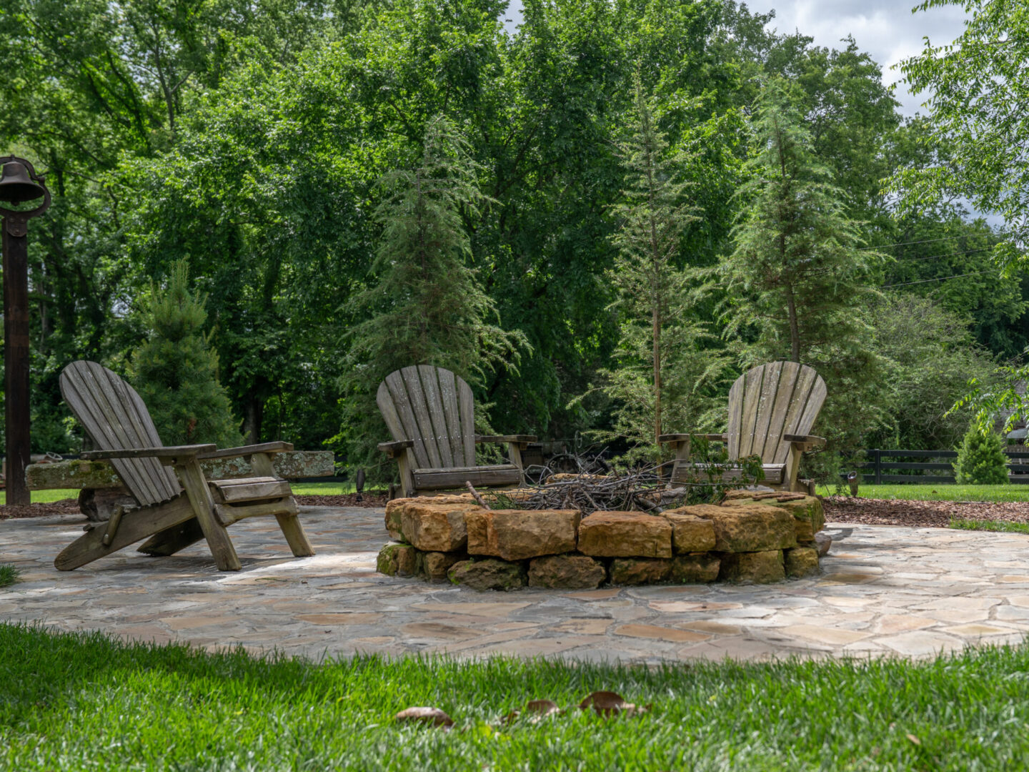 Outdoor fire pit area with wooden chairs on a stone patio surrounded by greenery.